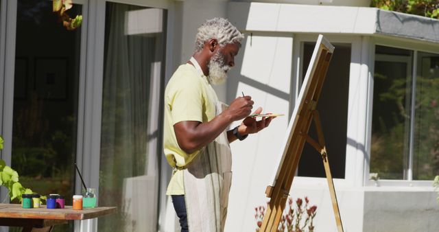 Senior man painting a canvas on an easel outside. Perfect for use in promotions related to senior hobbies, creativity, artistic pursuits, and relaxation. Ideal for illustrating outdoor leisure activities or art tutorials.