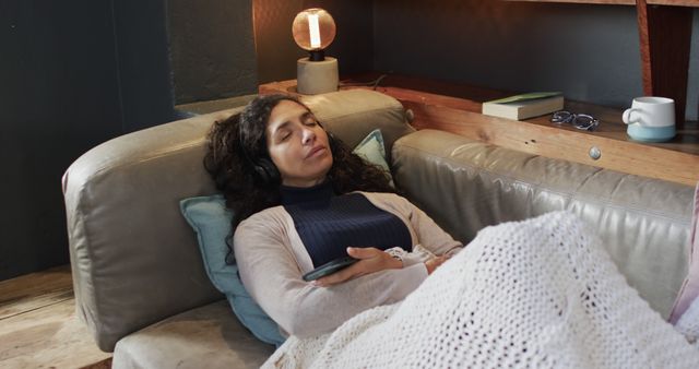 Woman lying on couch with headphones, listening to music and covered with a blanket. Ideal for content about relaxation, self-care, and lifestyle. Suitable for promotions of wellness, home living, and leisure products.