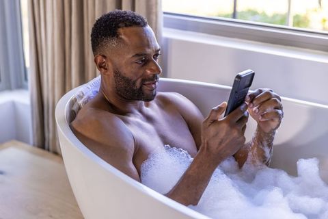 African american man relaxing in bubble bath using smartphone by sunlit window