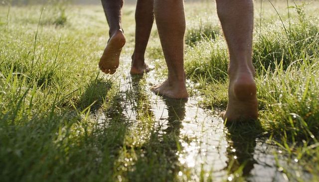 Barefoot adults walking through dewy grass trail splashing water in morning sunlight