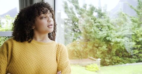 Woman gazing through window in mustard sweater, reflective home portrait with greenery