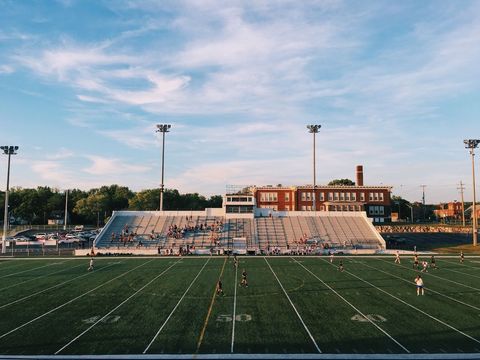 High school football stadium at sunset showing players on green turf and empty bleachers