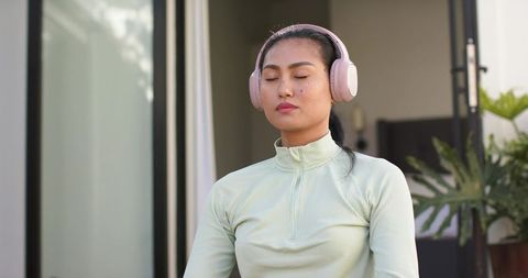 Mid-adult asian woman meditating on patio wearing pastel zip-up and pink headphones for mindfulness