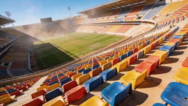 Empty multicolor stadium seats curving above sunlit grass field with sprinklers spraying