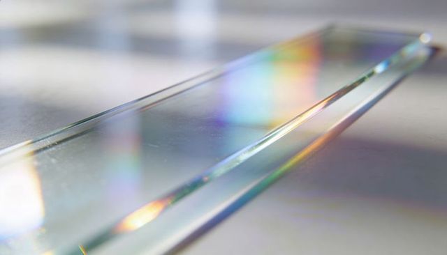 Long glass prism refracting rainbow light across pale tabletop macro with soft highlights