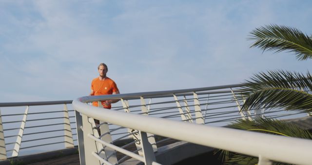 Man in orange shirt jogging on a bridge with railings and palm trees visible. This image is ideal for fitness and health-related advertisements, travel brochures, editorial content on maintaining a healthy lifestyle, and modern urban architecture promotions.