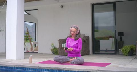 Mature woman practicing mindfulness on patio yoga mat beside pool using headphones