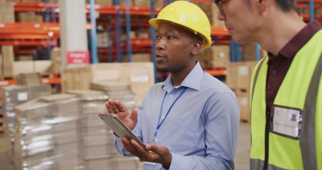 Warehouse manager discussing logistics with team member while holding a tablet in a distribution center. Workers wear safety equipment and discuss inventory management. Ideal for themes related to warehouse operations, logistics planning, or industrial teamwork.