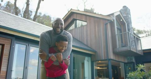 African american father laughing while giving son piggyback ride on modern wood-clad deck