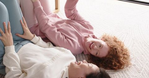 Smiling friends lounging on sunlit carpet wearing cozy pastel hoodies and leggings