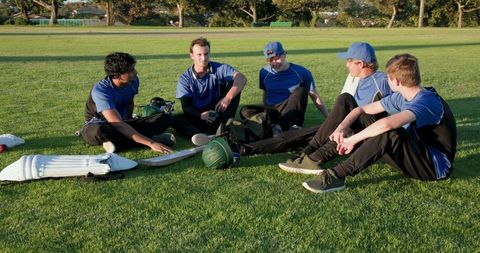 Young diverse cricket team sitting on grass sharing laughs and gear after training