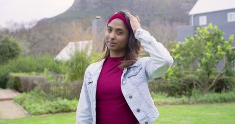 Indian woman adjusting burgundy headband in denim jacket standing in suburban garden