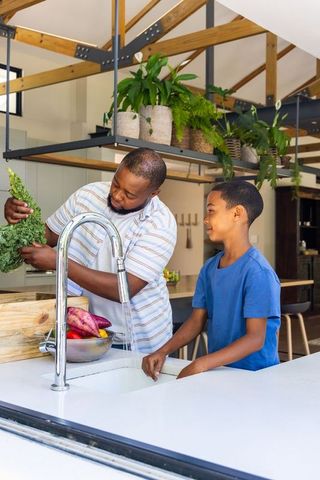 African american father and teen son washing greens in rustic kitchen family cooking