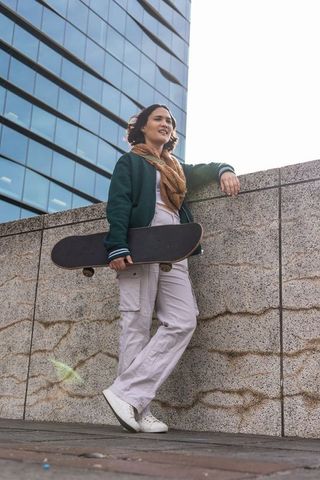 Young woman leaning on concrete wall holding skateboard in urban plaza, backlit streetwear