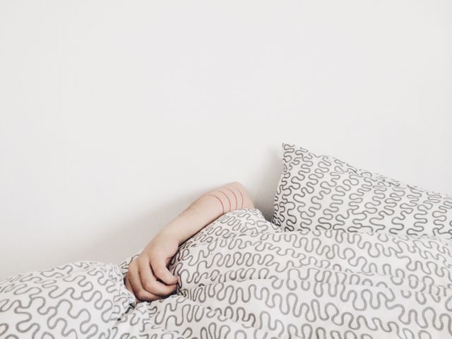 Person lying in bed, partially covered by a white quilt with a black, squiggly pattern. Only their arm is visible, extending out from under the quilt. The room features minimalistic design with a white wall. Ideal for promoting restful sleep, bedroom decor, minimalism, or a tranquil environment.