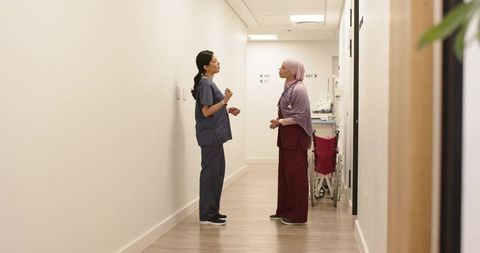 Diverse female clinicians talking in hospital corridor with wheelchair and stethoscope