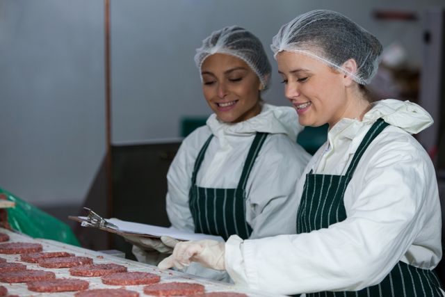 Two female butchers in a meat factory are maintaining records on a clipboard while wearing safety gear including hairnets, gloves, and uniforms. This image can be used for articles or advertisements related to the food industry, meat processing, quality control, workplace safety, and teamwork in industrial settings.