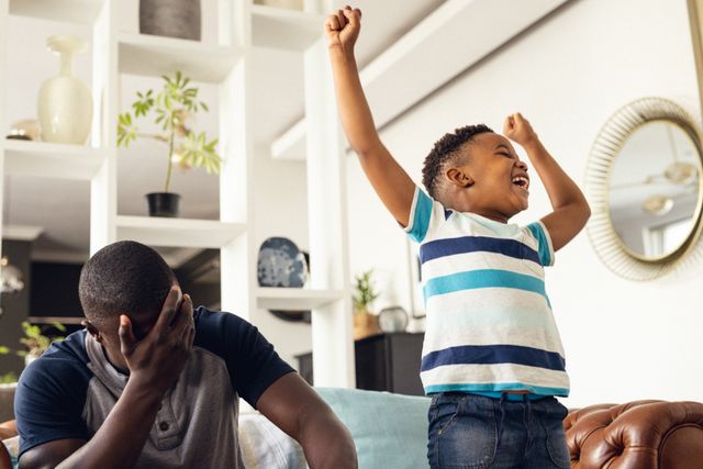 Happy african american son celebrating winning game with father in living room. Enjoying quality family time together at home.