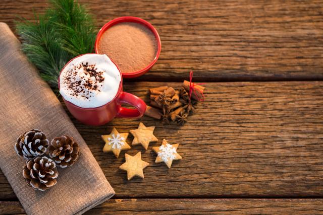 Perfect for holiday-themed promotions, this image captures a cozy Christmas setting with a red mug of hot chocolate topped with whipped cream, star-shaped cookies, pine cones, and cinnamon sticks on a rustic wooden table. Ideal for use in holiday greeting cards, festive blog posts, or seasonal advertisements.