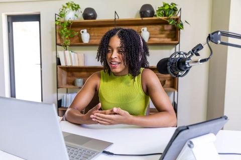 African woman podcasting from home studio speaking into broadcast microphone with laptop