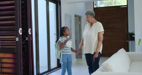 Black grandmother holding granddaughter hand in bright modern home doorway before school