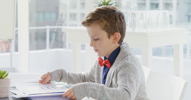 Young boy in a formal outfit reading documents in an office. He wears a gray cardigan and red bow tie, focusing intently. This image can be used for business training, educational materials about kids in professional settings, or promoting children's educational programs.
