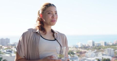 Woman standing on balcony holding glass cups on tray, gazing at ocean and city skyline