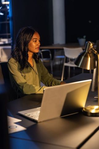 Asian woman working late at modern office desk with laptop under warm gold lamp