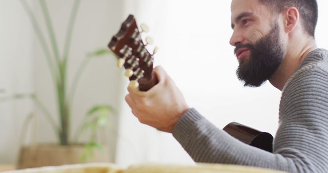 This image shows a man with a beard, sitting comfortably at home, smiling as he plays an acoustic guitar. The relaxed and casual atmosphere makes this perfect for illustrating concepts related to hobbies, leisure activities, music practice, enjoyment, and home life. It can be used in articles and advertisements that promote musical instruments, home entertainment, or personal time for hobbies.