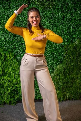 Smiling woman posing with raised arms near green hedge wearing mustard top and beige pants