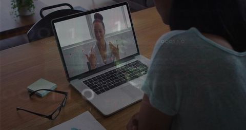 Woman having virtual meeting on laptop at home desk wearing patterned blouse, glasses