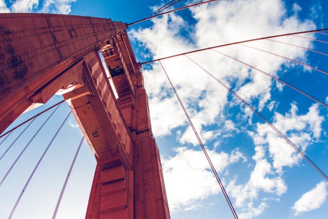 This striking image captures an upward perspective of a famous suspension bridge, set against a bright blue sky with scattered clouds. Ideal for use in travel brochures, architectural studies, tourism websites, and articles highlighting engineering marvels or infrastructure. The image evokes a sense of adventure and exploration, making it perfect for any content promoting travel destinations or celebrating modern structural design.