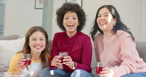 Diverse female friends enjoying cozy girls night in laughing on sofa with red drinks