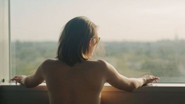 Young woman leaning on window sill gazing over urban skyline at sunrise with bare back
