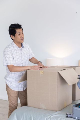 Asian man packing cardboard boxes on bed during apartment move, minimalist serene interior