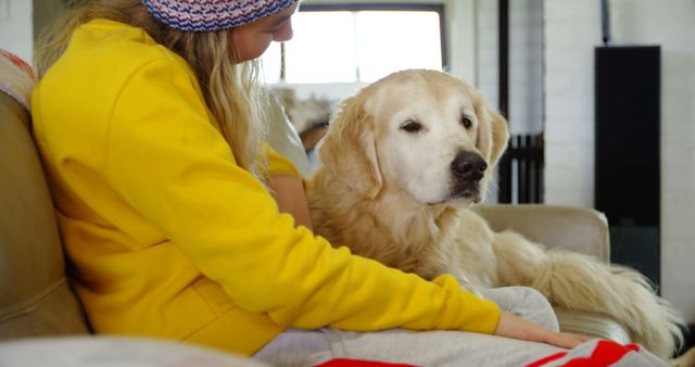 A woman in casual winter clothing sitting on a sofa with her golden retriever dog, showcasing a moment of relaxation and companionship in a cozy home environment. Ideal for use in promotional material for pet care products, home decor, or lifestyle blogs focusing on pet ownership and domestic comfort.