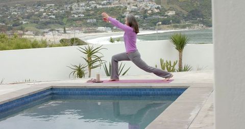 Mature woman practicing yoga lunge poolside on coastal terrace with succulents