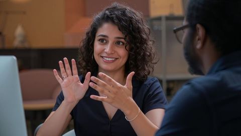 Curly-haired woman gesturing enthusiastically during collaborative office discussion over laptop
