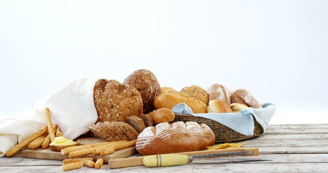 Wide variety of fresh, rustic bread and baked goods displayed on a wooden table. Includes different types of breads and crackers, placed next to a knife for cutting. Designed for uses in baking recipes, healthy eating articles and food blogs. Perfect for bakery advertisements, artisanal bread presentations and culinary promotions.