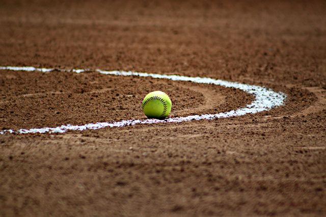 Softball resting on baseline of sandy field, highlighting smooth, curved lines in background. Ideal for sports-related content, athletic endorsements, team introductions, and game announcements.