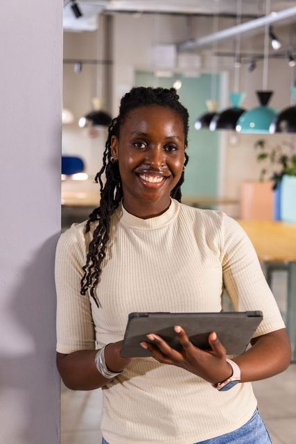 African american woman leaning on column smiling while using tablet in coworking space