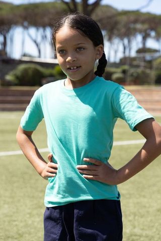 Confident young girl standing hands on hips on sports field in teal shirt and navy shorts