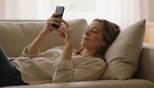 Reclining woman using smartphone on beige sofa relaxing at home scrolling messages