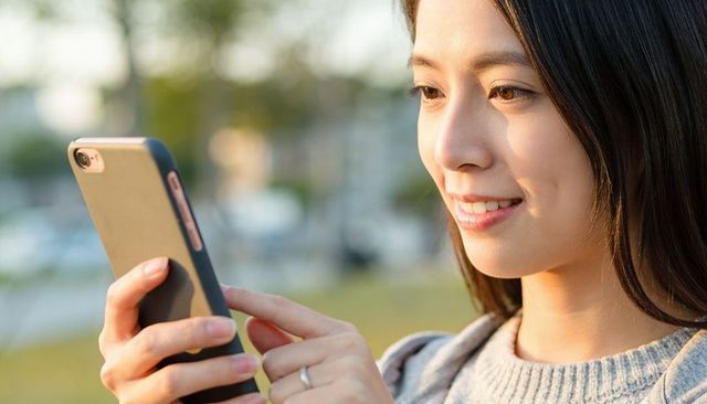 Smiling young asian woman tapping smartphone in park during golden hour, outdoor lifestyle