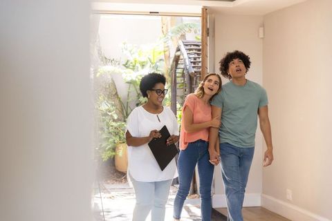 Realtor showing sunlit home entry to young couple, touring courtyard with leafy stairs