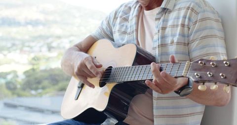 Adult man fingerpicking acoustic guitar by window with city view and wristwatch