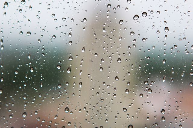 Clear raindrops sitting on window glass with an out-of-focus background, suggesting a rainy day. Can use for illustrating weather conditions, capturing mood or ambiance, nature details, or metaphors for clarity/obscurity, isolation or reflections.