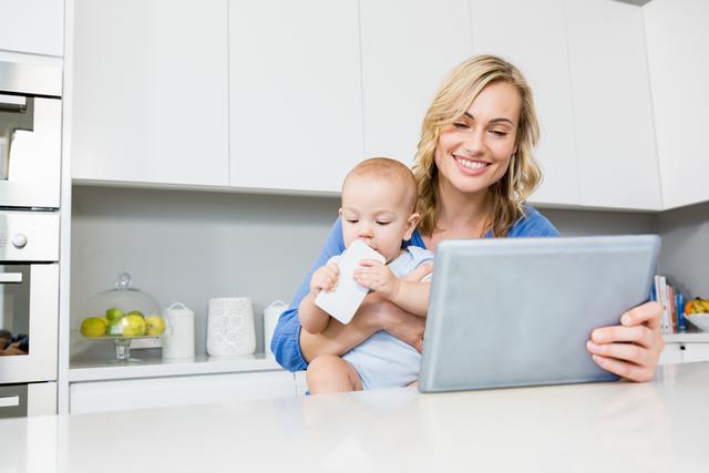 Mother holding baby boy while using a digital tablet in a modern kitchen. The baby is drinking from a bottle while the mother is smiling and engaged with the tablet. This image can be used for topics related to modern parenting, multitasking, family life, technology in the home, and convenience in daily routines.