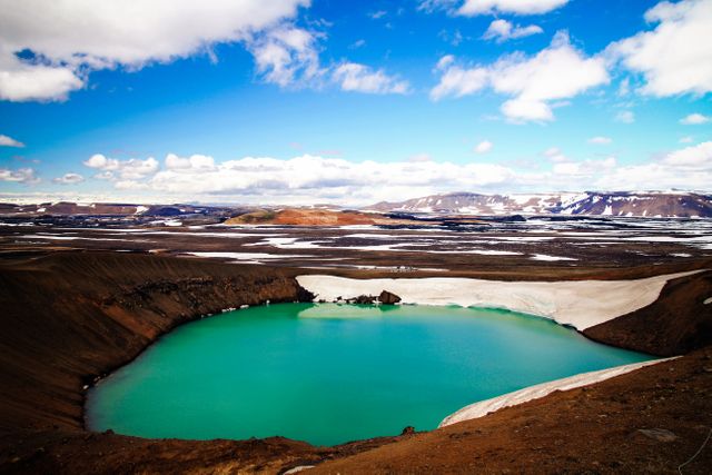 Panoramic view of a stunning blue crater lake surrounded by a volcanic landscape with snowy patches and a clear sky. Perfect for travel brochures, nature and adventure-themed articles, destination promotions, and environmental studies.