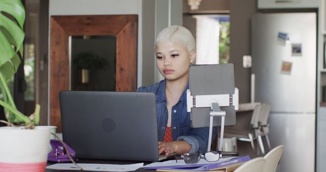 Young woman with short blonde hair working remotely from home using a laptop. Workspace includes modern tablet stand, plant, and documents. Great for illustrating concepts of remote work, modern technology, home offices, and personal productivity.